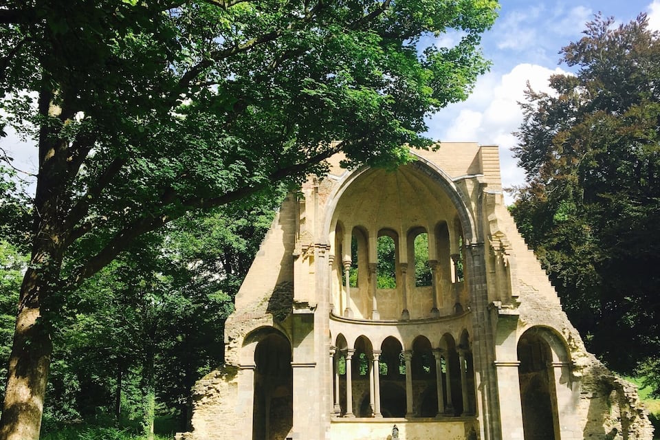 Ein historisches Steinhaus mit Rundbogenfenstern und einem zentralen Eingang, umgeben von üppigen Grünpflanzen und Gras.