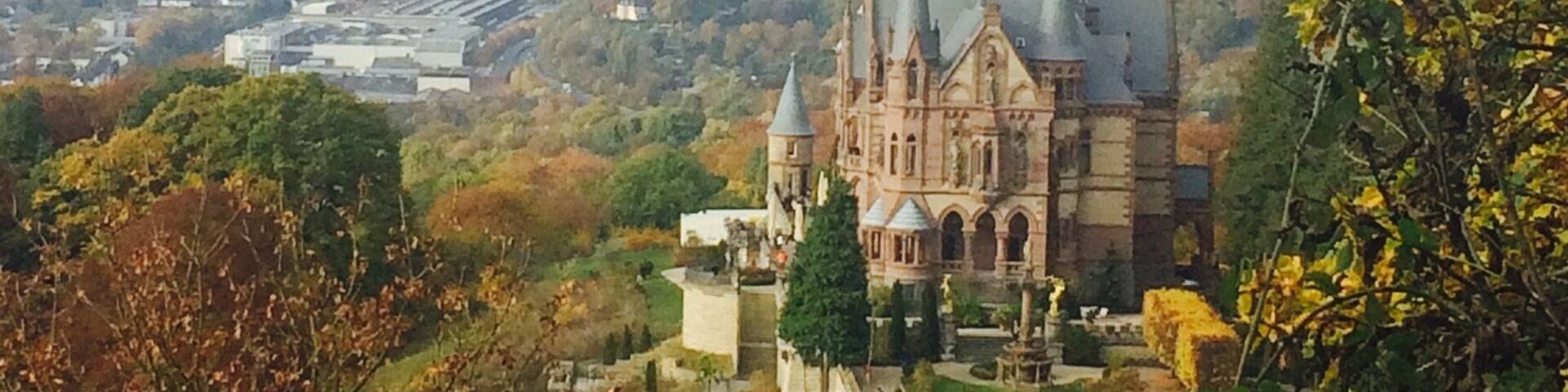 Schloss Drachenberg and view of Bonn beyond. Schloss Drachenburg is a private villa in palace style constructed in the late 19th century. It was completed in only two years (1882–84) on the Drachenfels hill in Königswinter, a German town on the Rhine near the city of Bonn. Baron Stephan von Sarter (1833–1902), a broker and banker, planned to live there, but never did.
Today the Palace is in the possession of the State Foundation of North Rhine-Westphalia. It is served by an intermediate station on the Drachenfels Railway. You can ride the train or hike from the bottom.