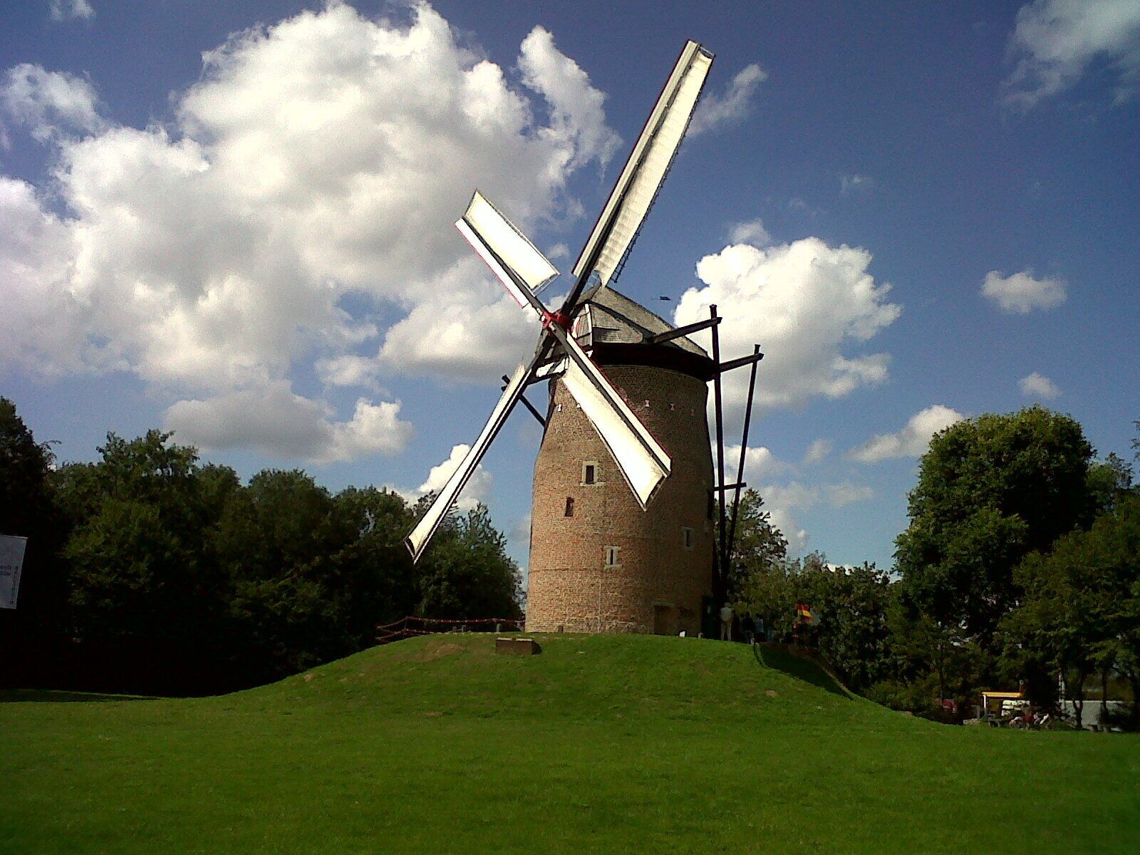 A traditional windmill with large sails, set against a backdrop of a clear blue sky and lush green trees.