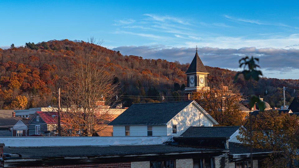 A town with a prominent church steeple, houses, and a hill with autumn foliage.