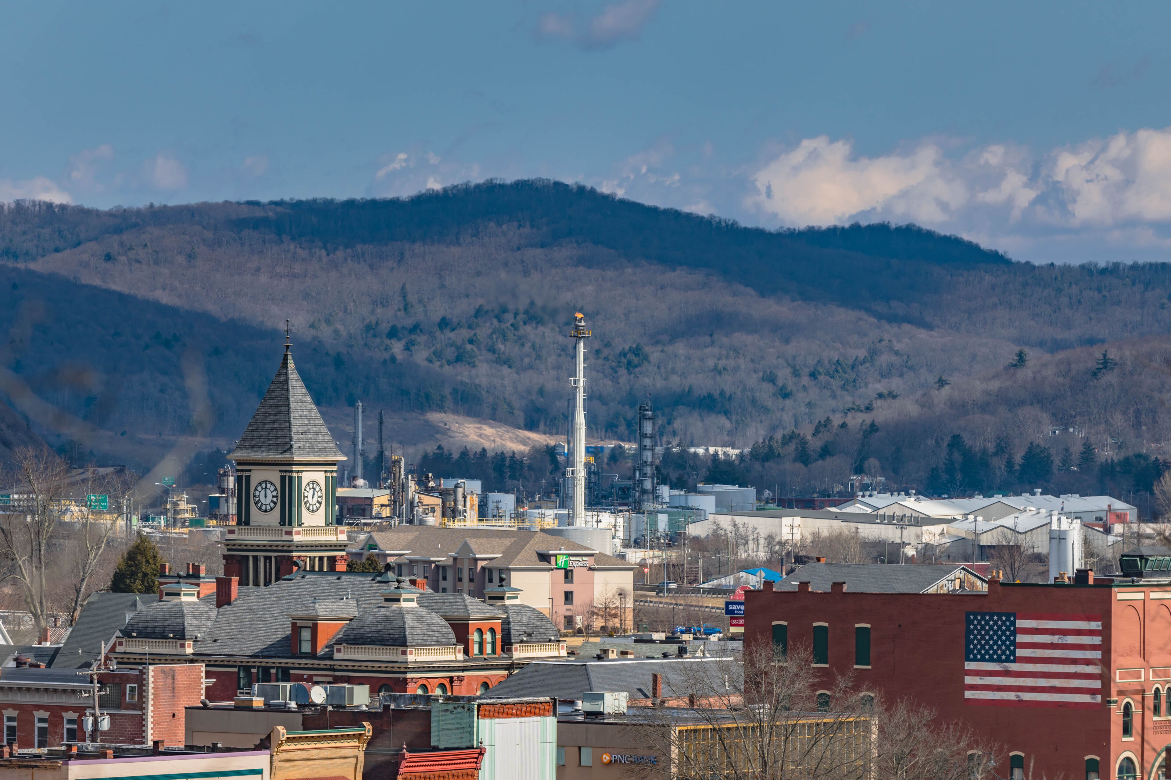 A town with a prominent clock tower, a large American flag, and a backdrop of mountains.