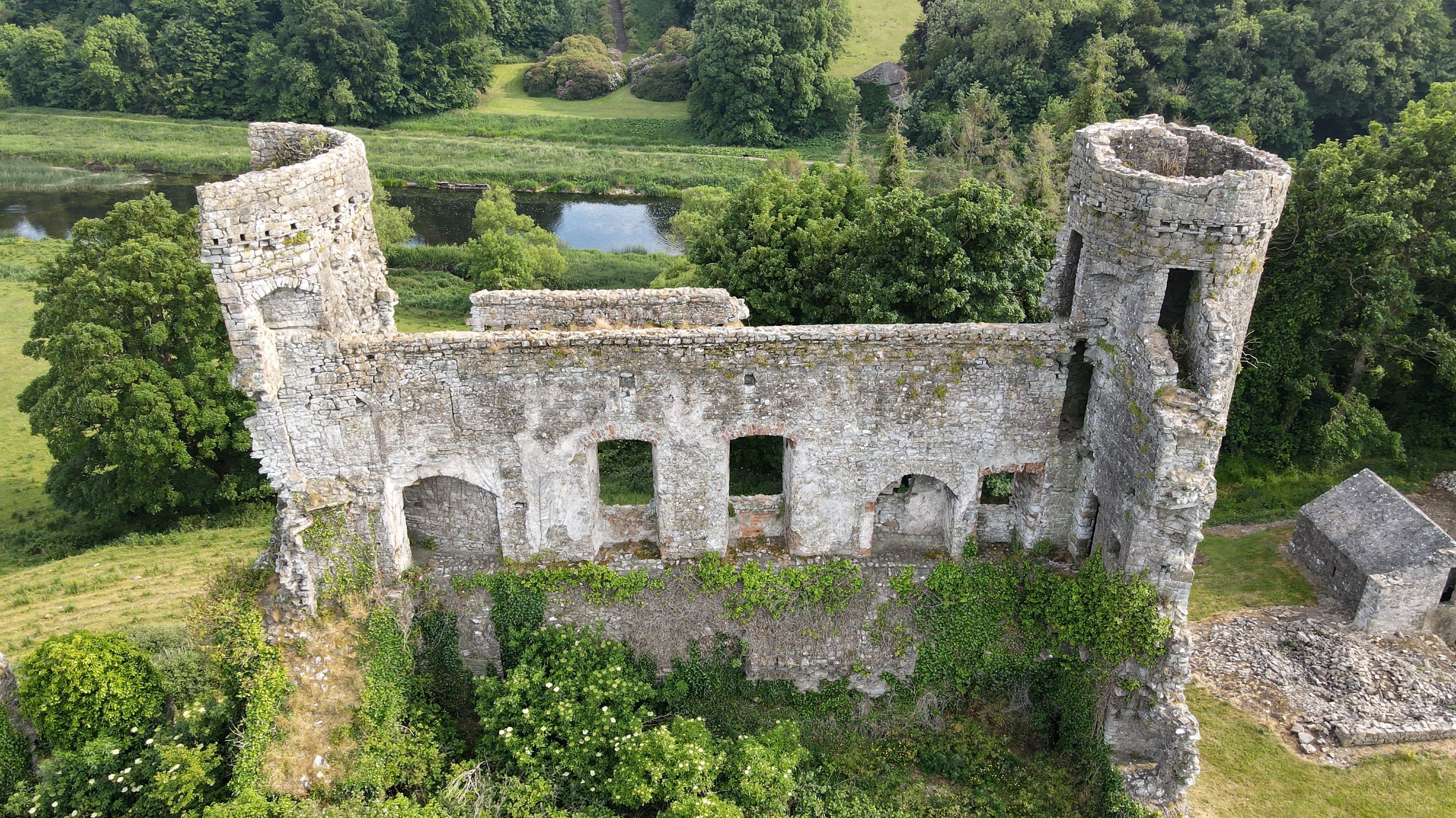 An aerial view of a historic stone castle surrounded by lush greenery and a small pond.