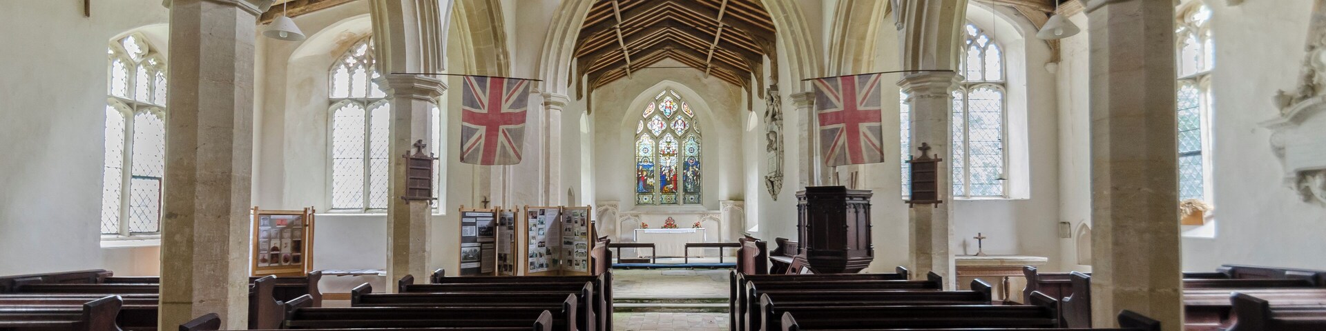 Interior de una iglesia con techos abovedados, bancadas de madera, vidrieras y banderas británicas.