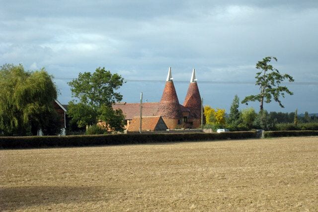A rural landscape with a brick building featuring two conical structures, surrounded by fields and trees.