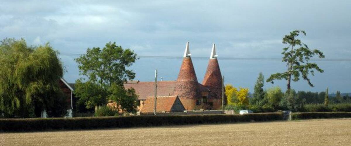 A rural landscape with a brick building featuring two conical structures, surrounded by fields and trees.