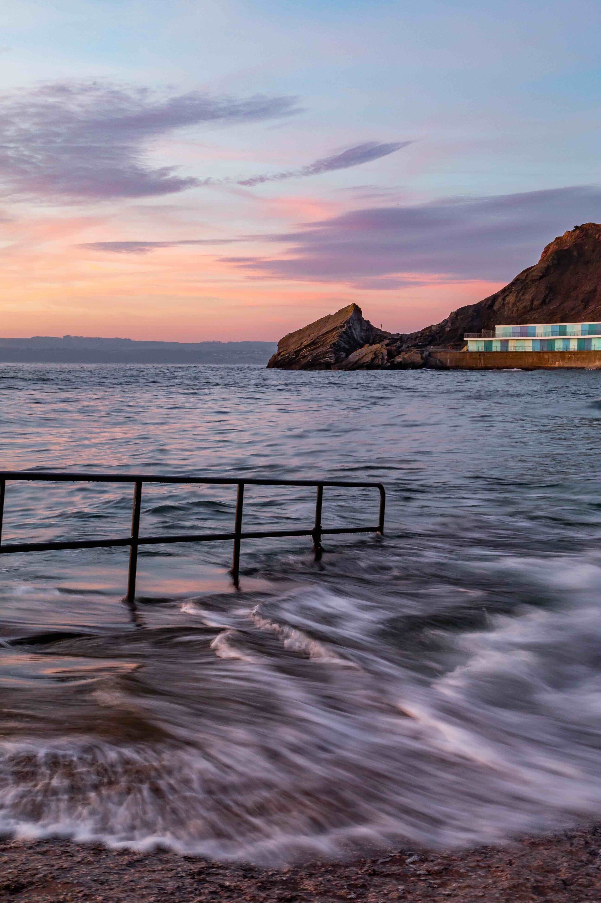Una puesta de sol costera con olas, un promontorio rocoso y un edificio junto al mar.