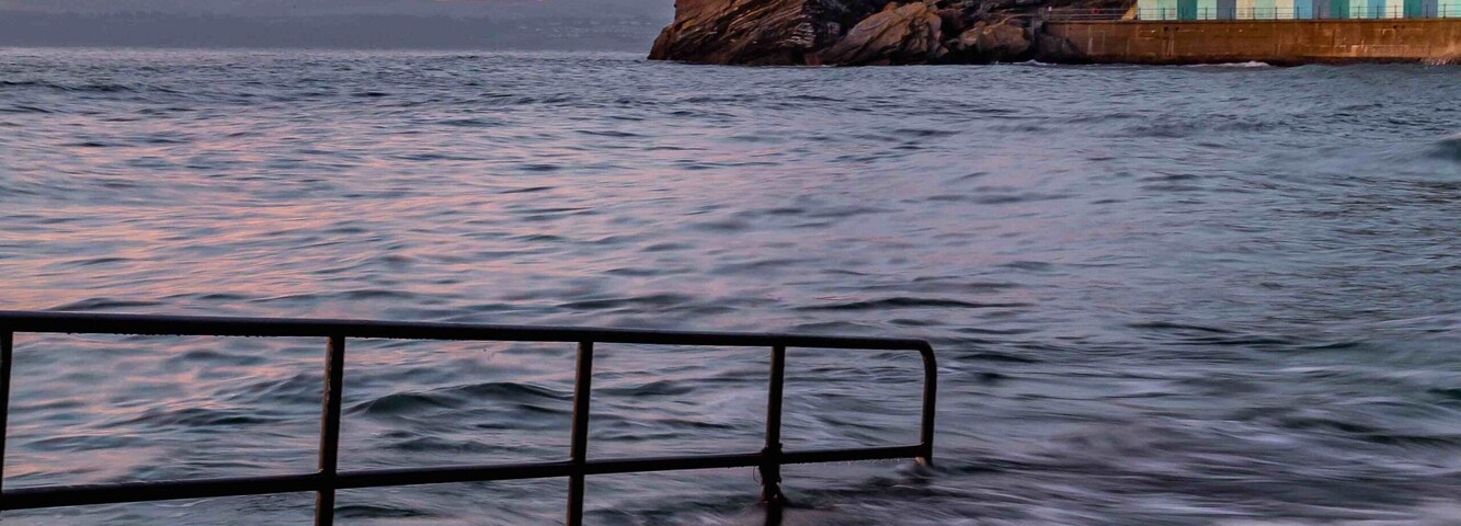 Una puesta de sol costera con olas, un promontorio rocoso y un edificio junto al mar.