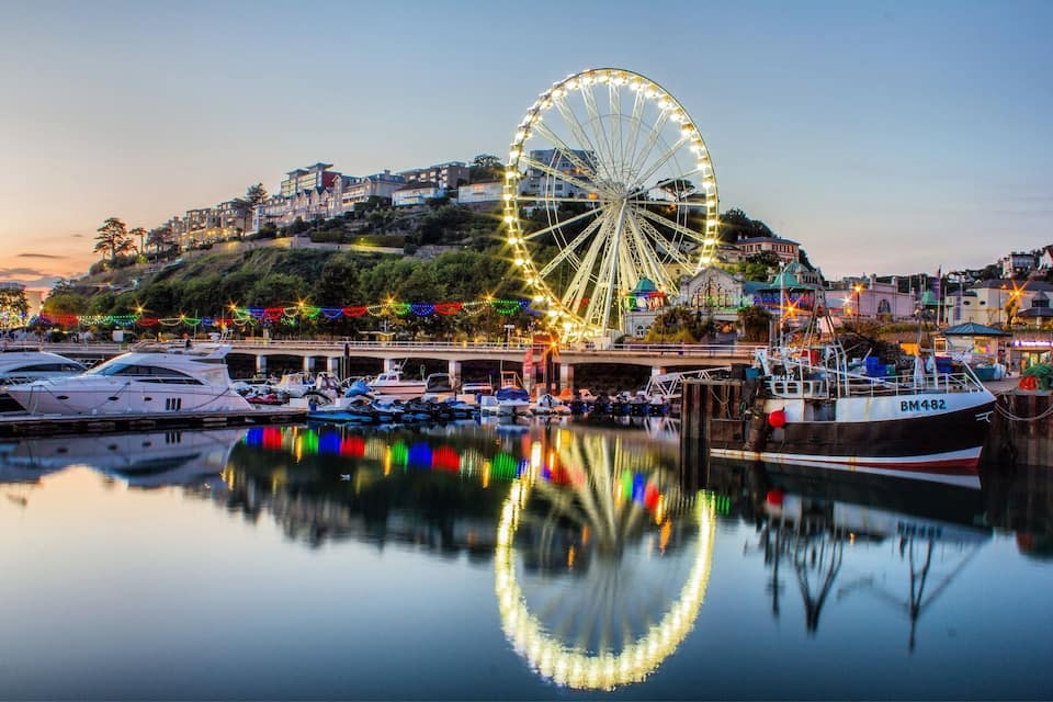 A waterfront marina with a large Ferris wheel, boats, and colorful lights reflecting on the water.