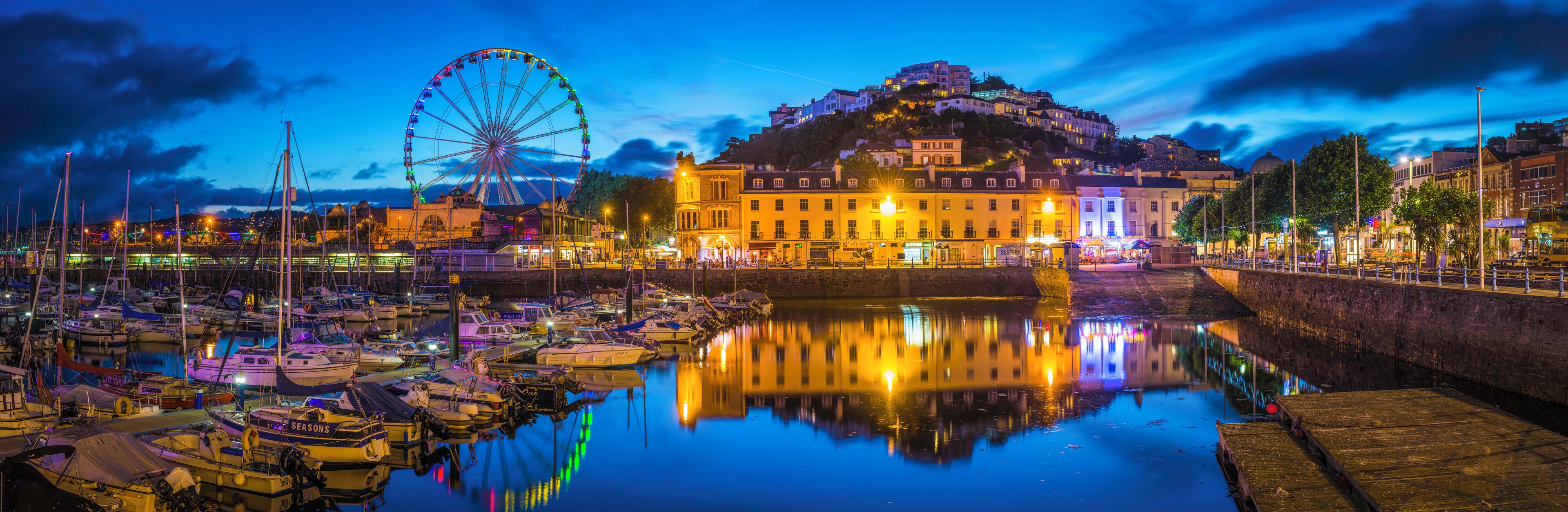 The hotels and villas, quayside shops, pubs and restaurants of Torquay overlooking the harbour illuminated at dusk.