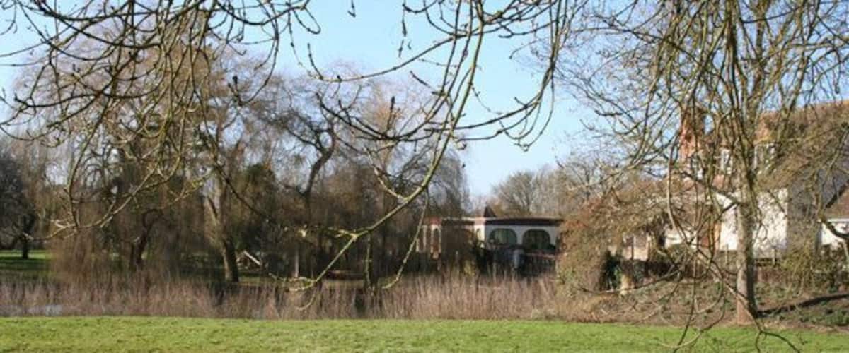 A grassy field with a bridge, trees, and houses in the background.