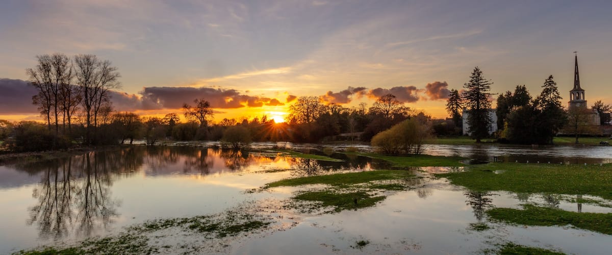 A sunset over a calm lake with reflections, surrounded by trees and a church tower.