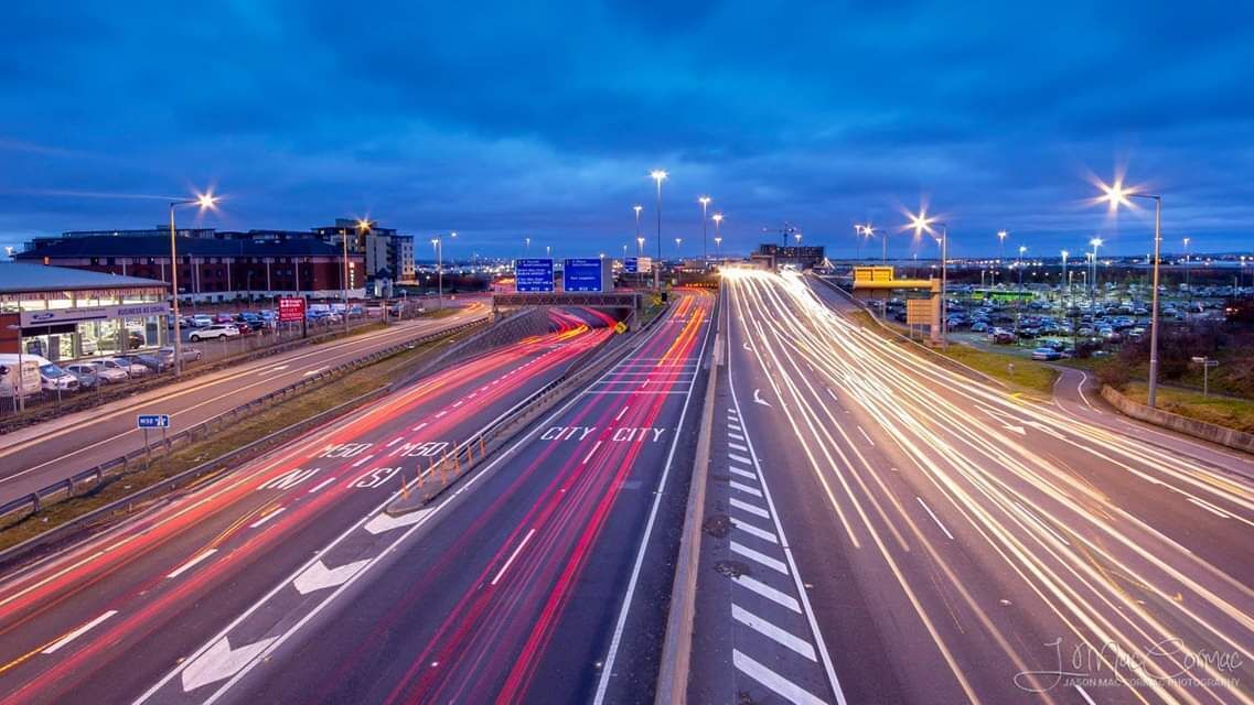 A multi-lane highway with light trails, a parking lot, and a building in the background.
