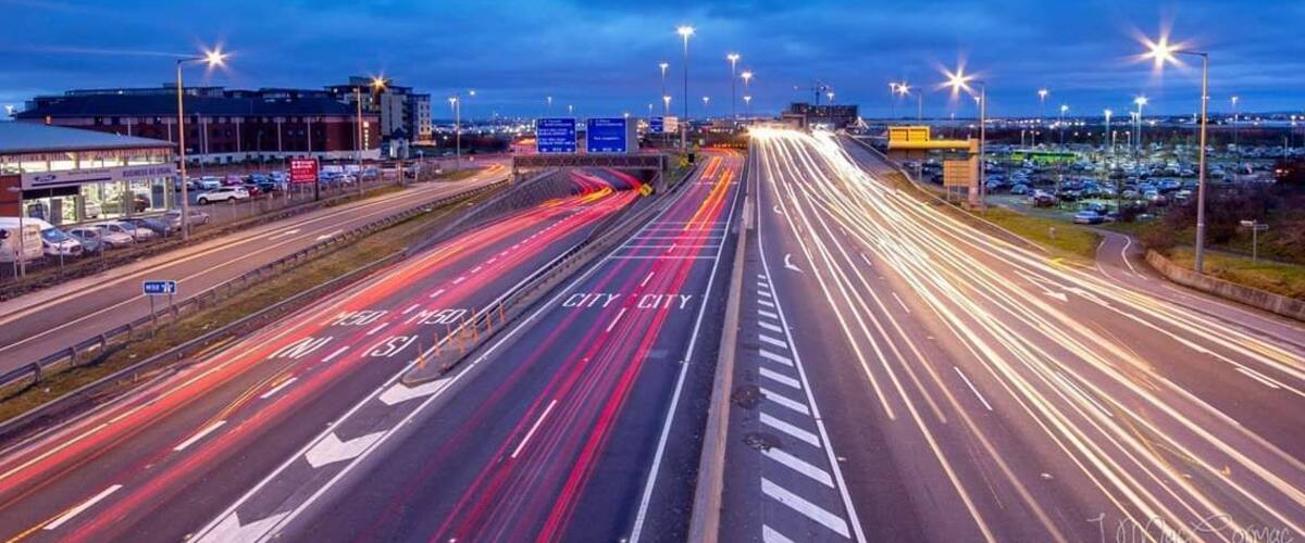 A multi-lane highway with light trails, a parking lot, and a building in the background.