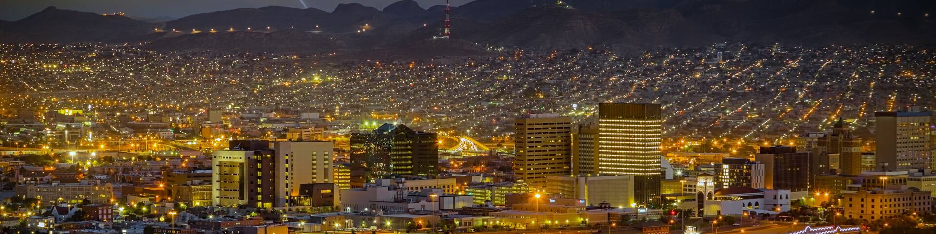 A cityscape at night with a lightning strike in the distance and mountains in the background.