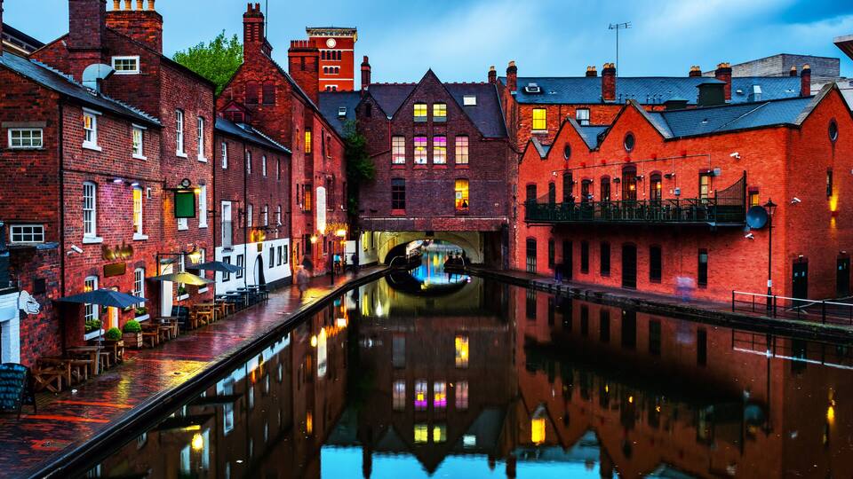 A canal with buildings reflecting on the water, a boat, and a red brick structure with a clock tower.