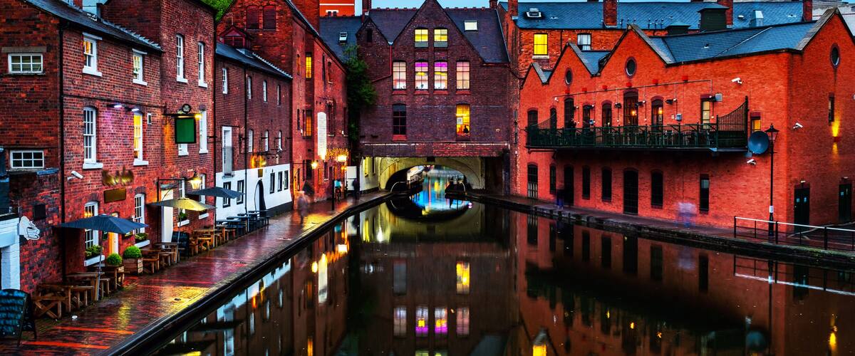 A canal with buildings reflecting on the water, a boat, and a red brick structure with a clock tower.