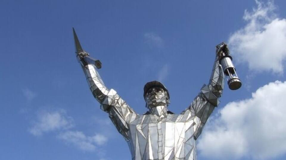 A metallic statue of a person holding a telescope and a book against a blue sky.