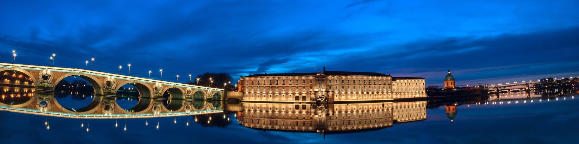 Un bâtiment historique doté d’un grand dôme, un pont aux arches circulaires se reflétant dans l’eau, et une vue sur la ville en arrière-plan.