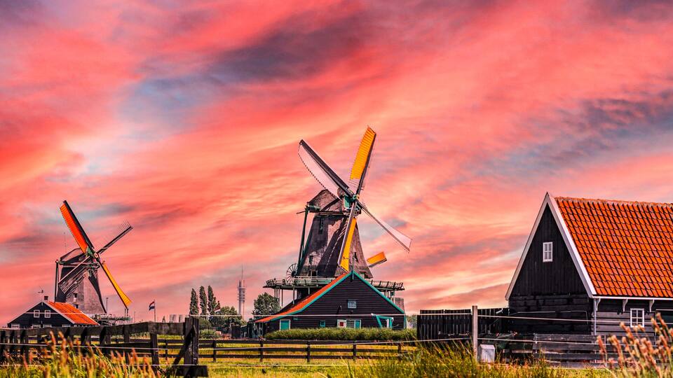 Windmills, a wooden house with a red roof, and a field under a colorful sunset sky.