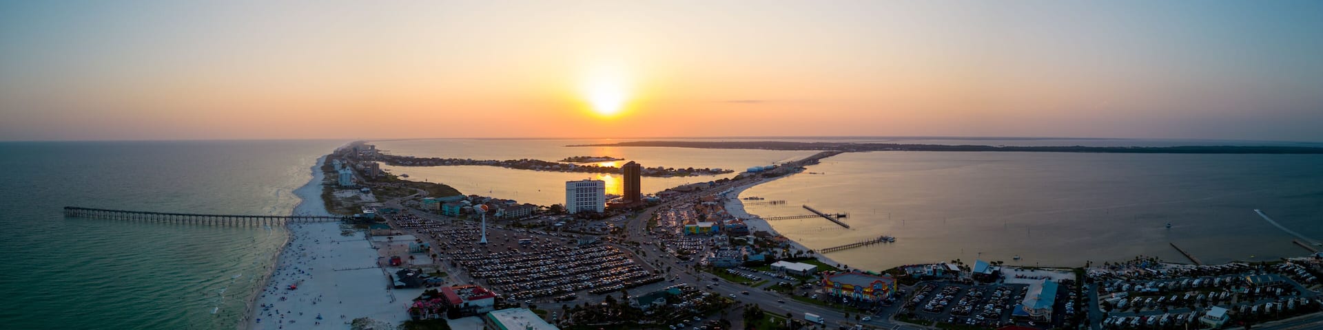 A coastal cityscape at sunset with a prominent pier extending into the water, a dense urban area with buildings, and a clear view of the ocean.