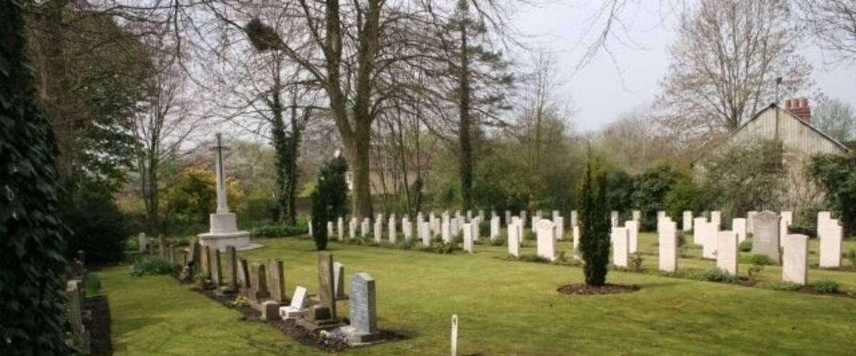 A cemetery with rows of headstones, a central monument, and a building in the background.