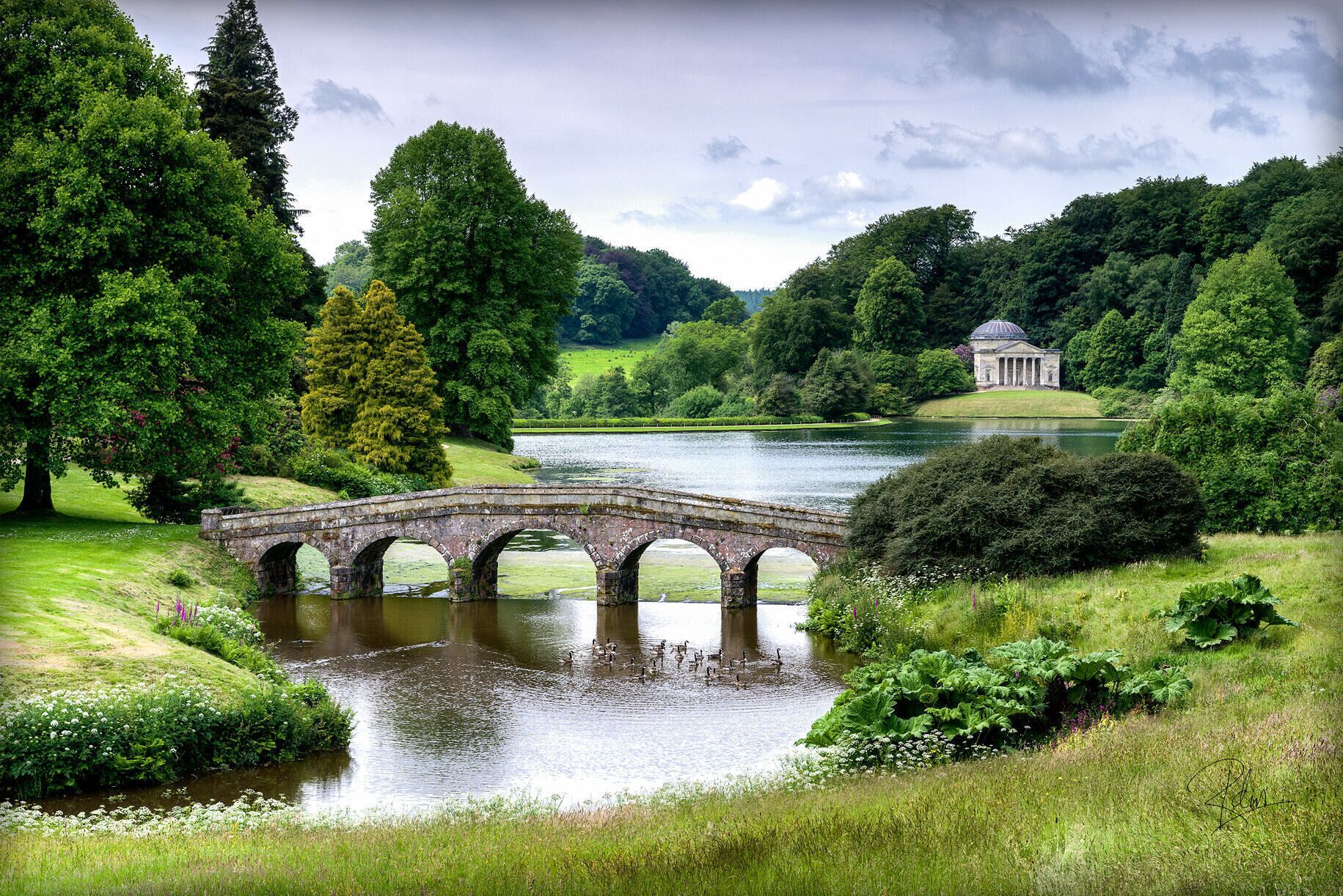A stone bridge over a calm river, surrounded by lush greenery and a classical building in the background.