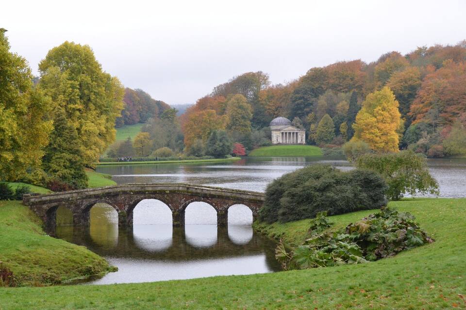 A stone bridge over a calm lake, surrounded by autumn-colored trees and a classical building in the background.
