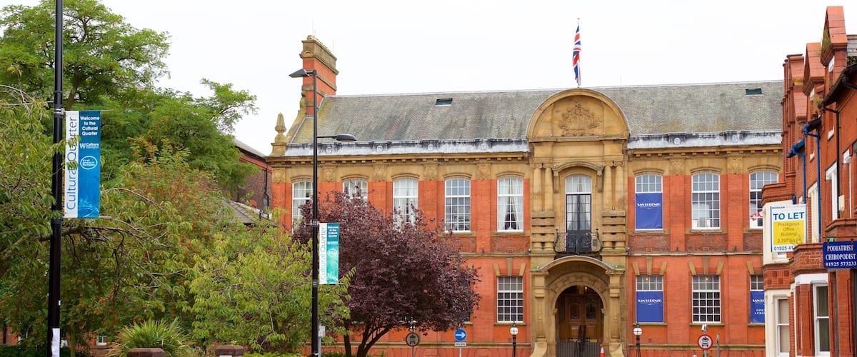A historic building with a red brick facade, a prominent entrance with a large archway, and a flag on top. There are cars parked in front, and a street sign indicating a one-way street.