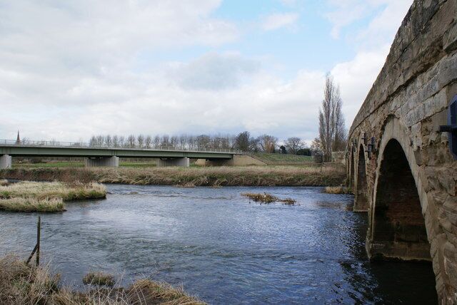 A stone bridge over a river, surrounded by grassy banks and a cloudy sky.