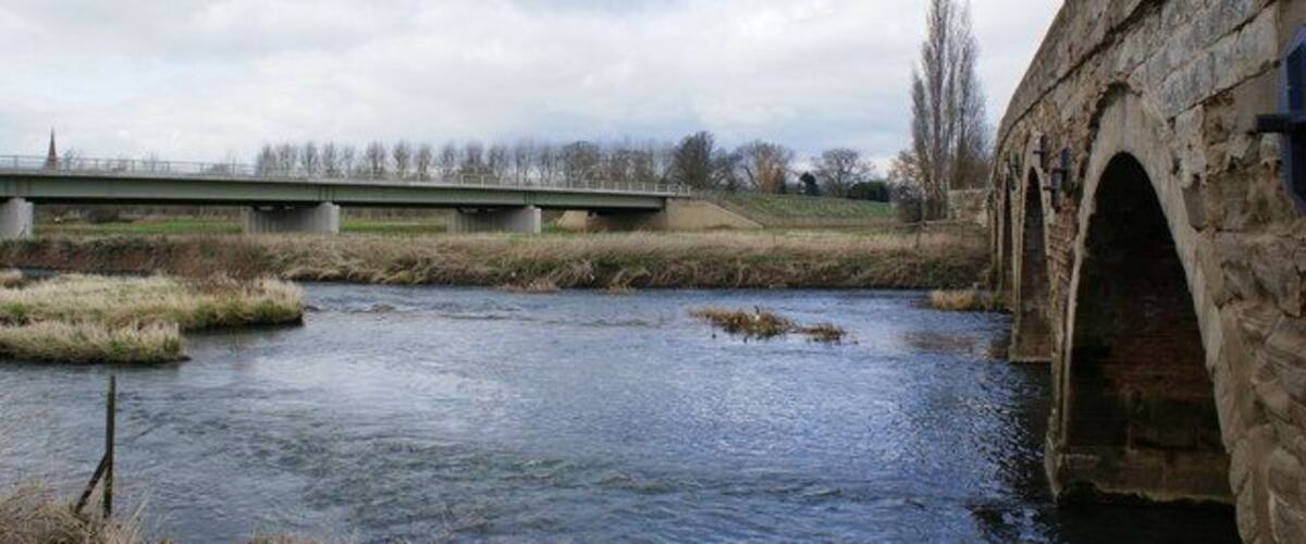 A stone bridge over a river, surrounded by grassy banks and a cloudy sky.