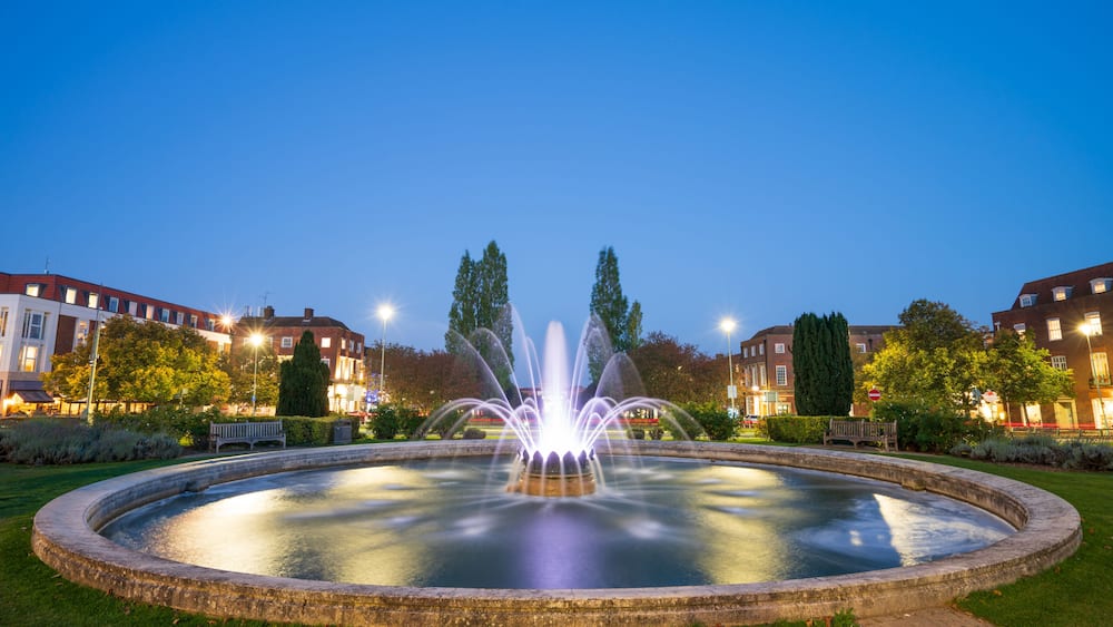 Une fontaine se trouve au centre d’un bassin circulaire, dont les jets d’eau s’élèvent au-dessus de l’eau. Elle est entourée de pelouse et d’arbres, et des bâtiments se dressent à l’arrière-plan.