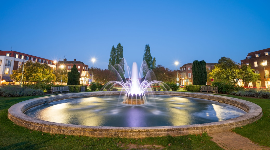 A fountain in a circular pool with water jets, surrounded by grass and trees, with buildings in the background.