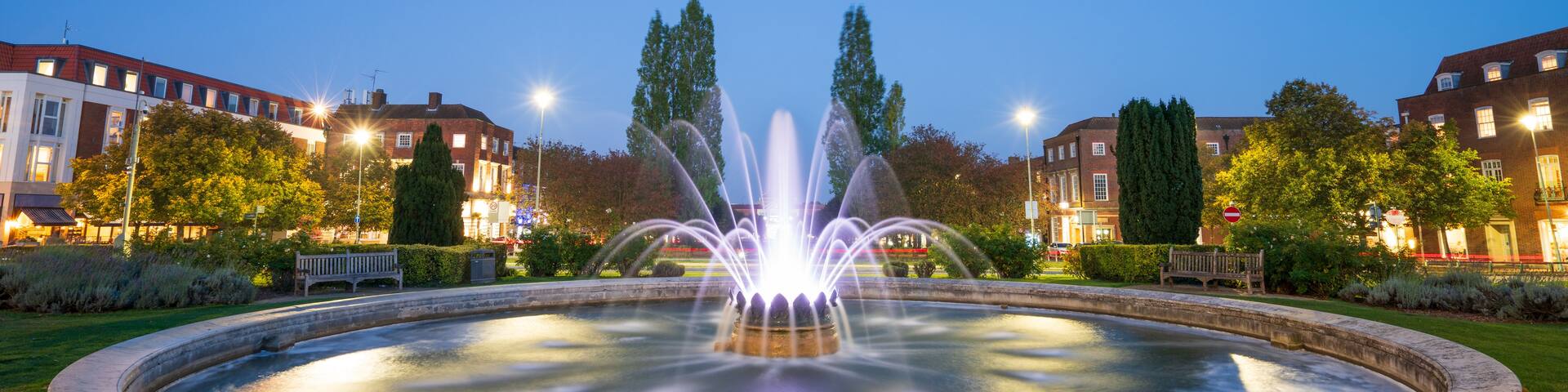 A fountain in a circular pool with water jets, surrounded by grass and trees, with buildings in the background.