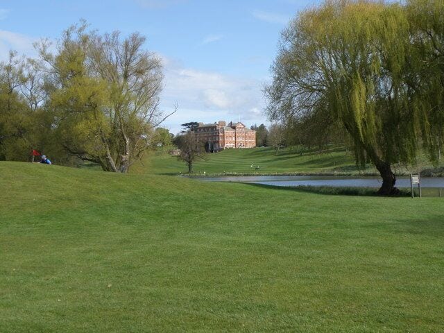 A grassy park with a pond, trees, and a building in the background.