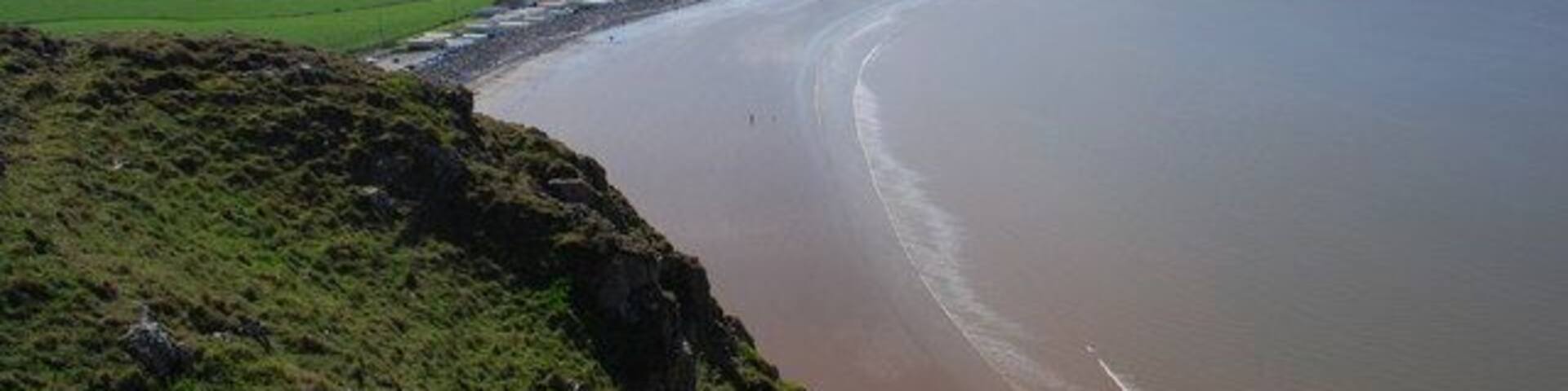 A coastal landscape with a wide sandy beach, grassy hills, and a clear blue sky.