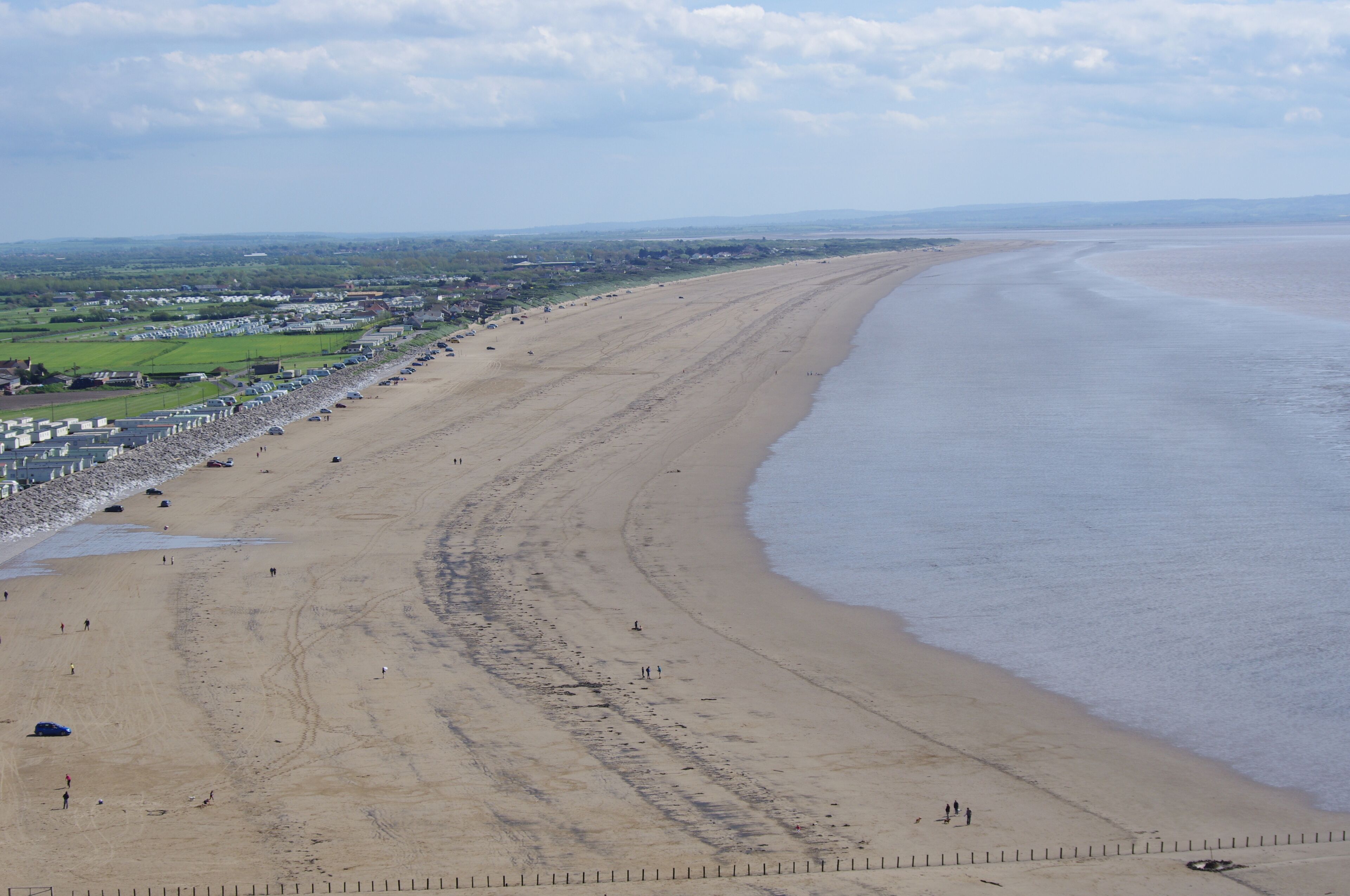 A wide sandy beach with a gentle curve, a fence line, and a distant view of houses and fields.