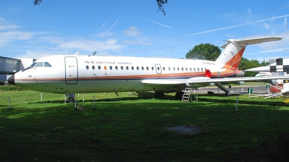 A white and orange airplane with "Fly By Light Control technology" on its side, parked on grass with a clear sky in the background.