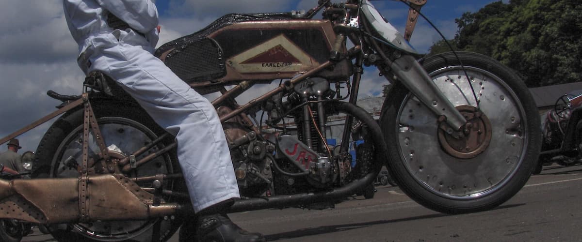 A vintage motorcycle with a large front wheel and a sidecar, ridden by a person in white protective gear.