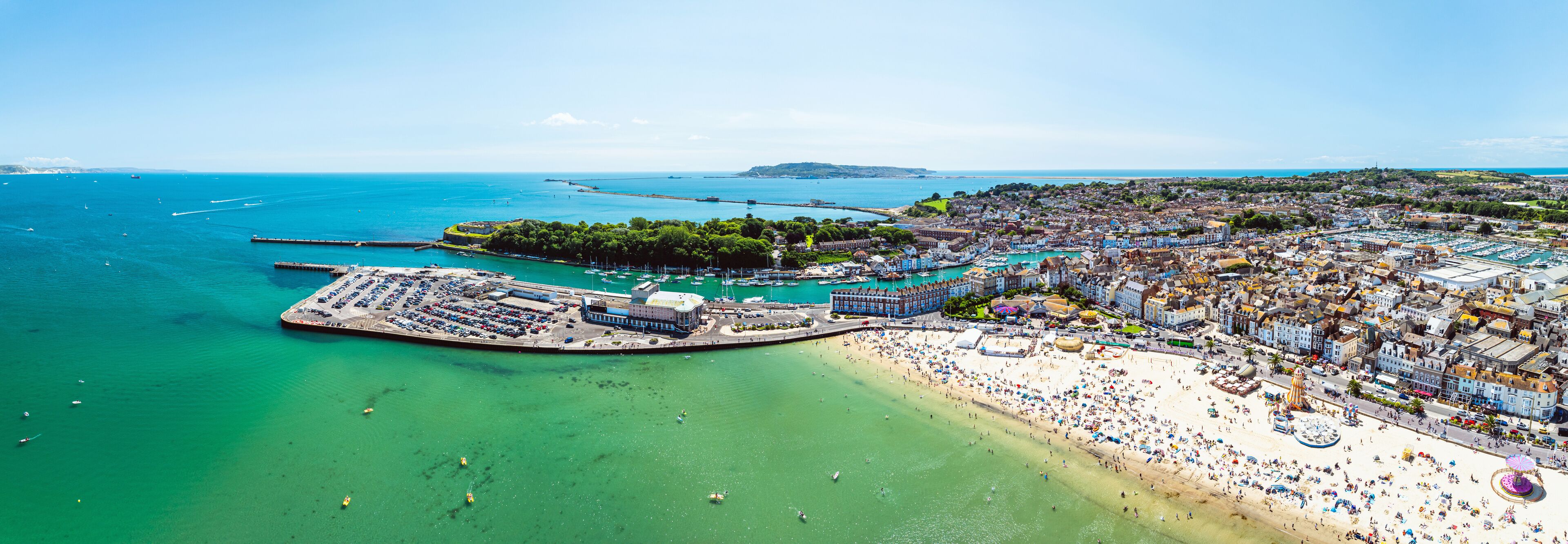An aerial view of a coastal town with a large beach, marina, and surrounding hills.