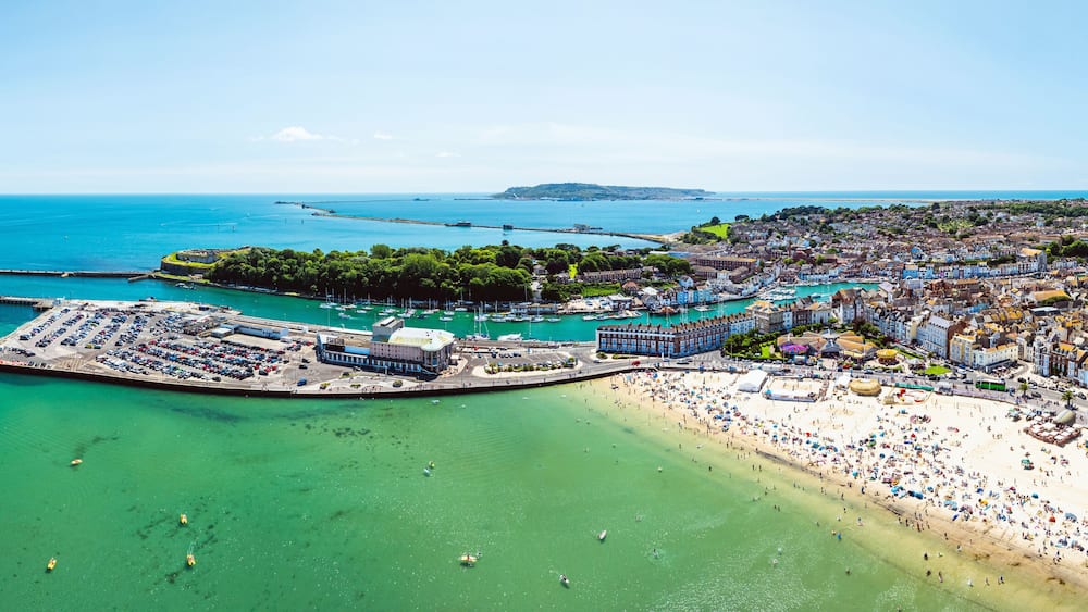 Eine Luftaufnahme einer Küstenstadt mit einem großen Strand, einer Marina und umliegenden Hügeln.