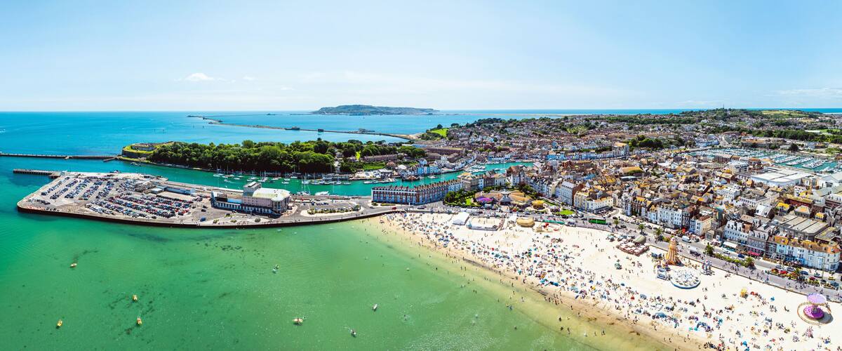 Vue aérienne d’une ville côtière avec une grande plage, un port de plaisance et les collines environnantes.