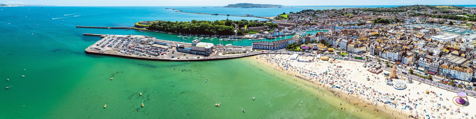 An aerial view of a coastal town with a large beach, marina, and surrounding hills.