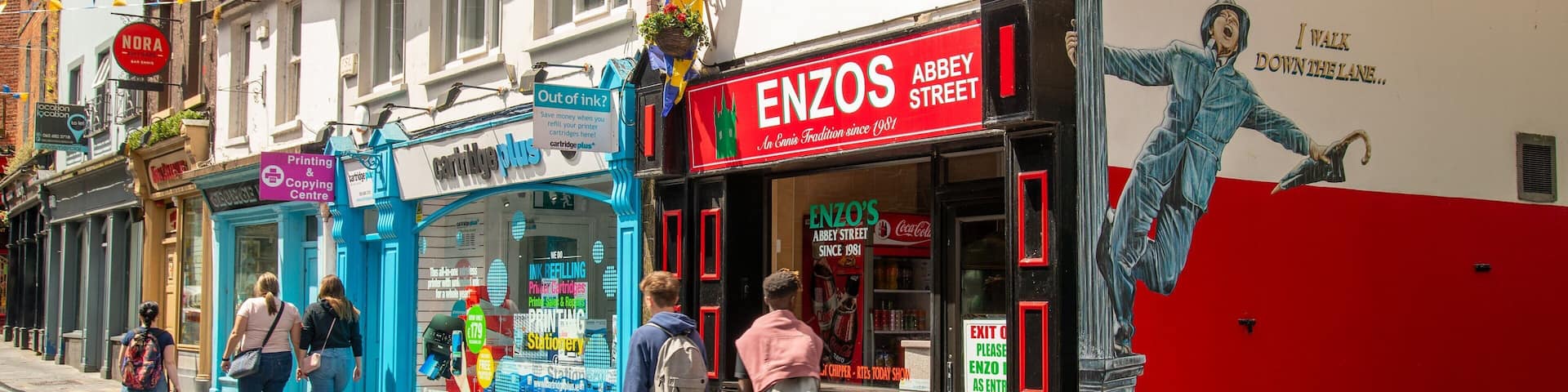 A street with shops, including ENZOS on Abbey Street, a printing and copying center, and a blue storefront.