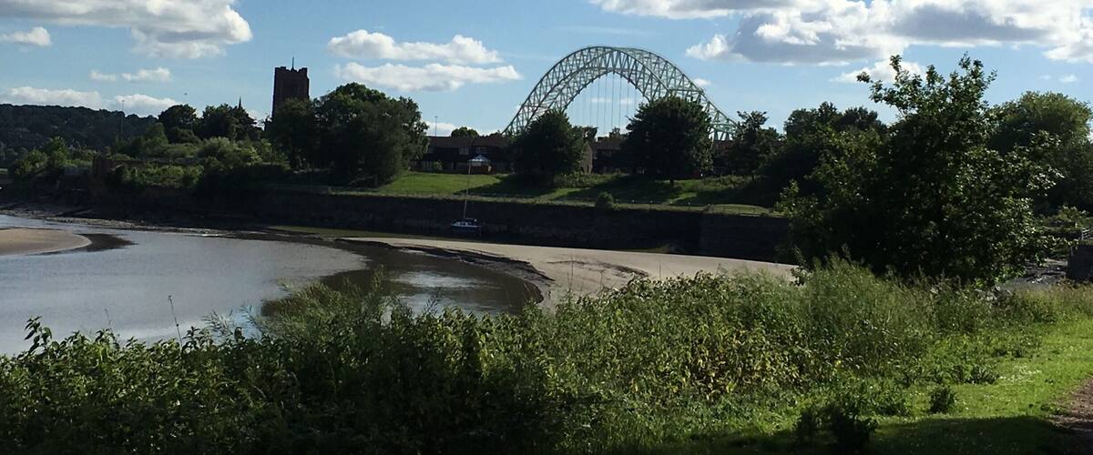 A coastal landscape with a large green bridge, a river, and a castle in the background.