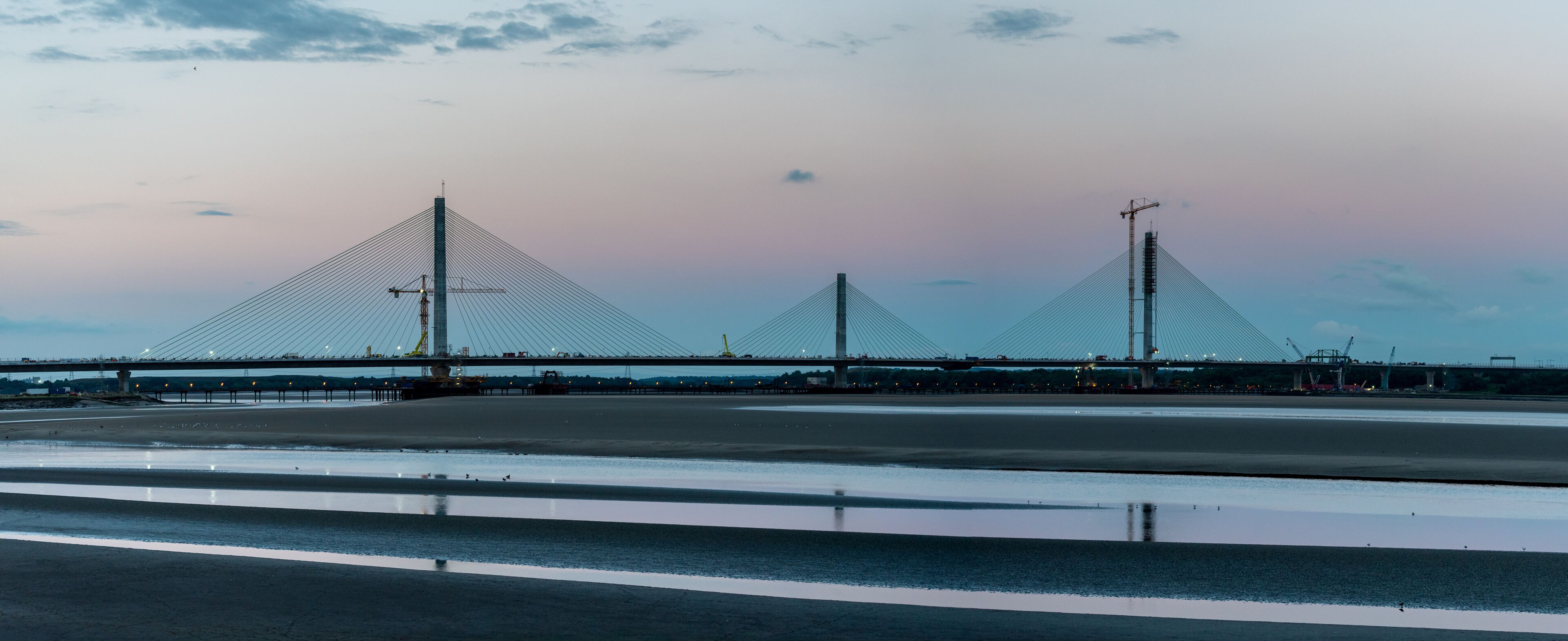A large suspension bridge with three towers, a calm river, and a sunset sky.
