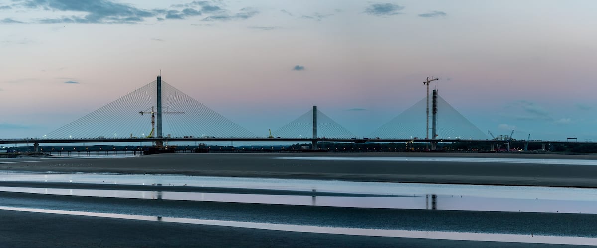 A large suspension bridge with three towers, a calm river, and a sunset sky.