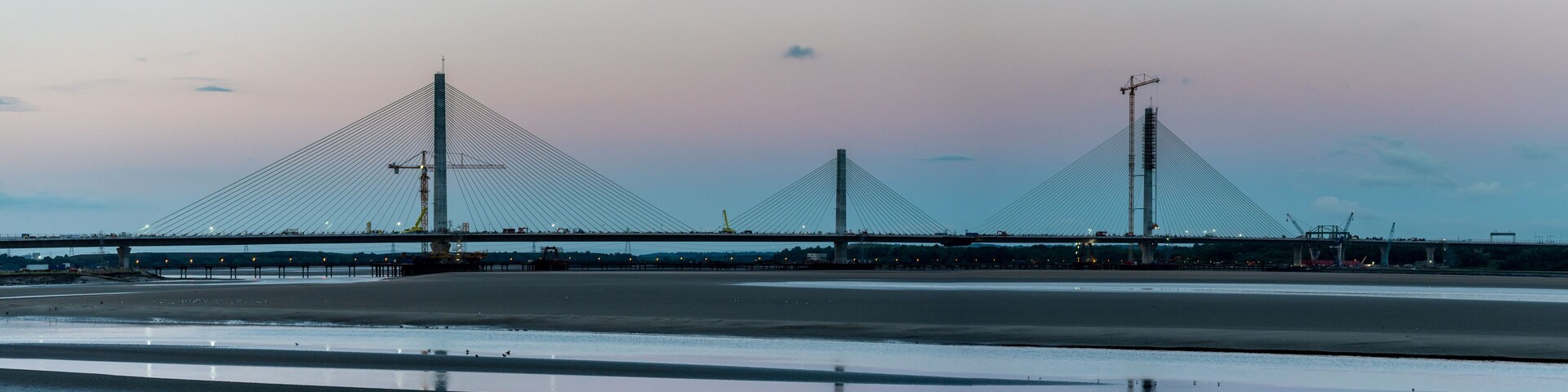 A large suspension bridge with three towers, a calm river, and a sunset sky.