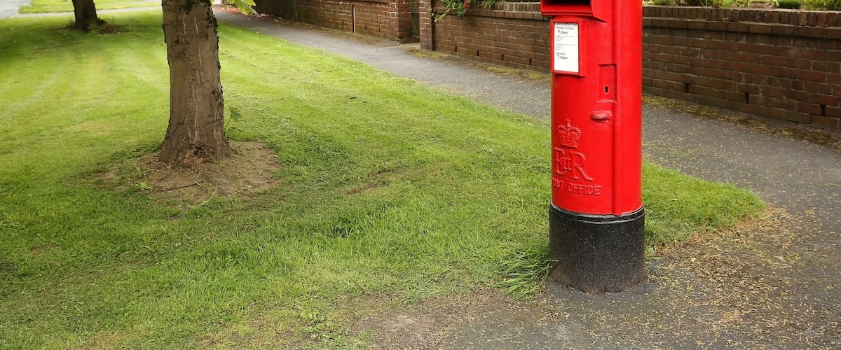 A red post box on a grassy path with a tree and brick wall in the background.