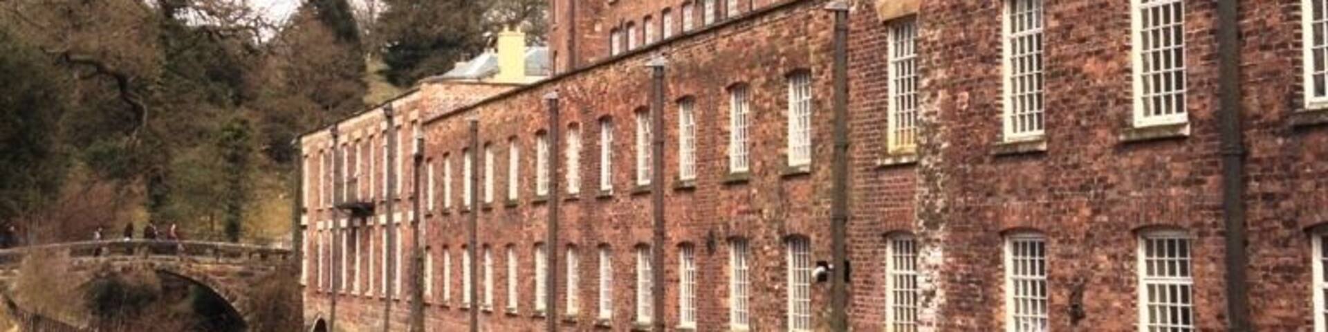 A large brick building with multiple windows and a chimney, adjacent to a canal with a bridge.