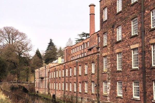 Un grand bâtiment en briques, doté de nombreuses fenêtres et d’une cheminée, situé à côté d’un canal qu’enjambe un pont.